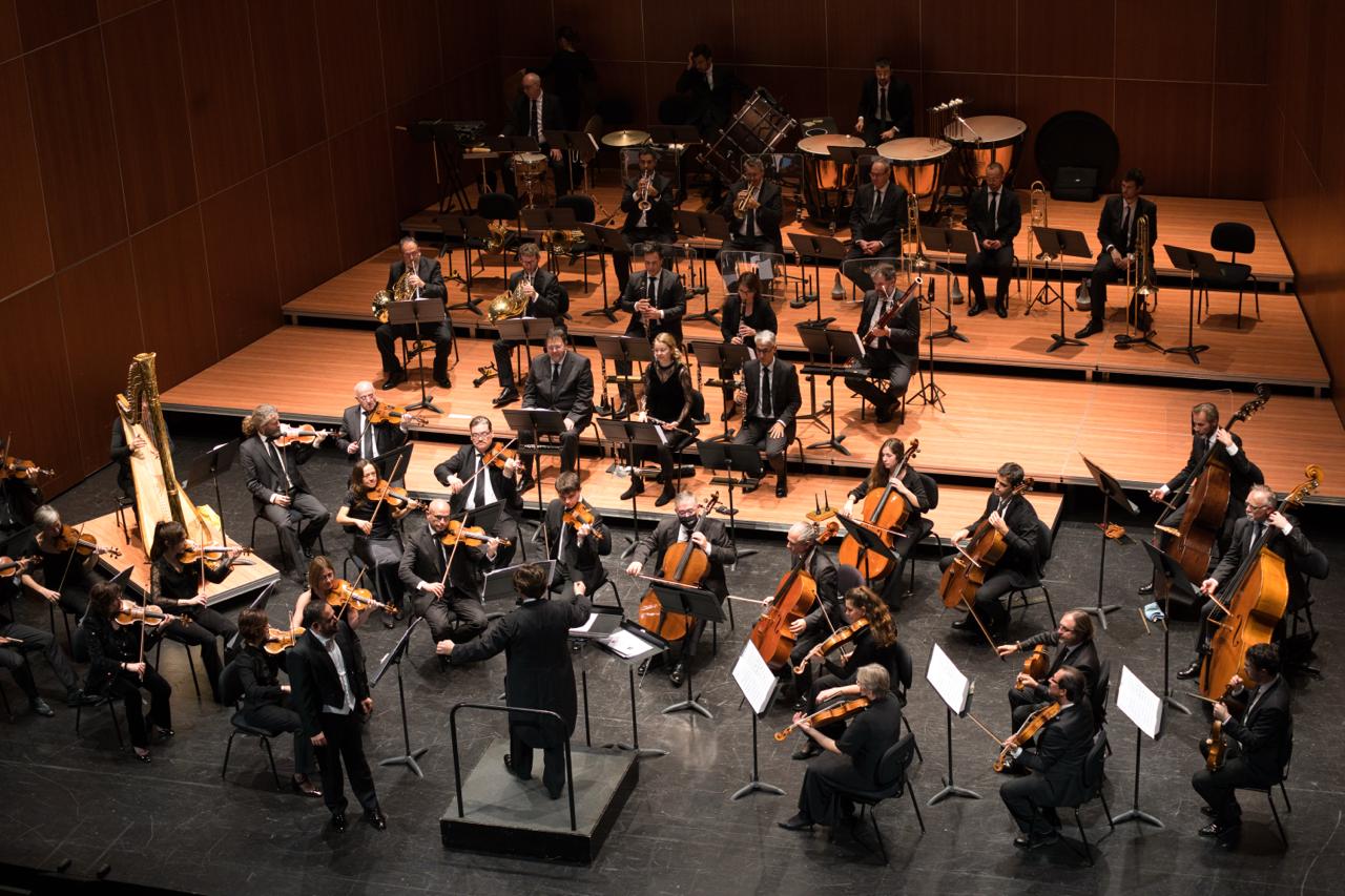Músics de l’Orquestra Simfònica Illes Balears tocant instruments i el director al centre en un escenari de concert.